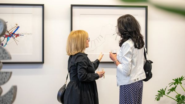 Two women discuss abstract art at a gallery while holding drinks.