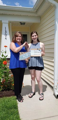Two women celebrating outside a newly sold house with signs and keys.