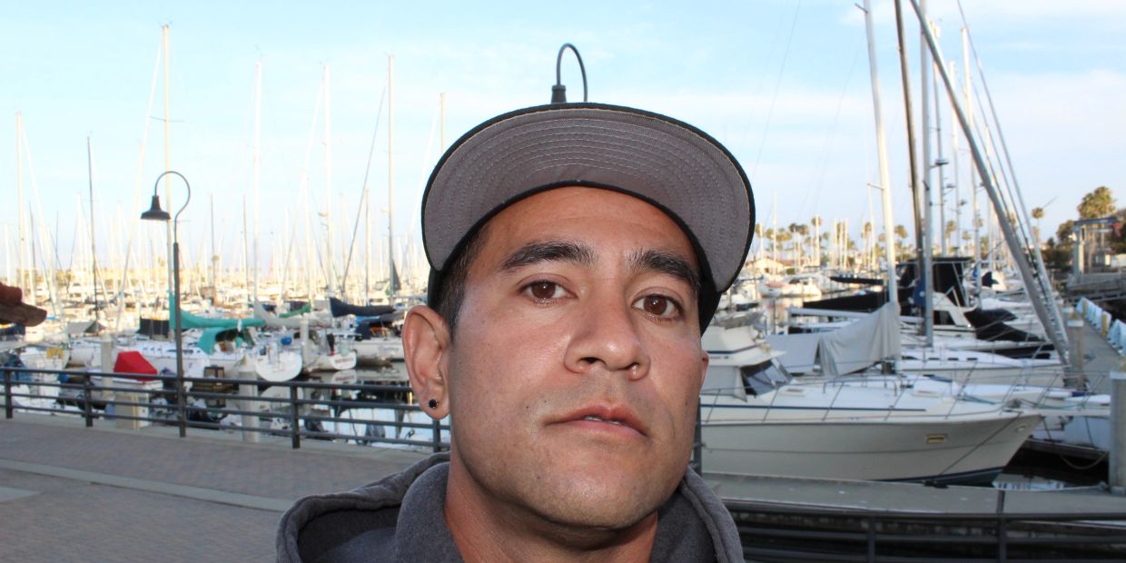 A man wearing a cap poses near a marina with numerous boats.