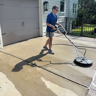 A man pressure washes a driveway in front of a house on a sunny day.