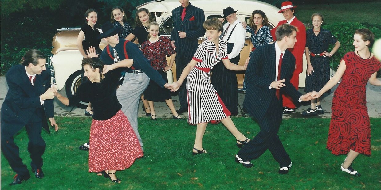 Group dancing in vintage attire on grass with classic car in background.