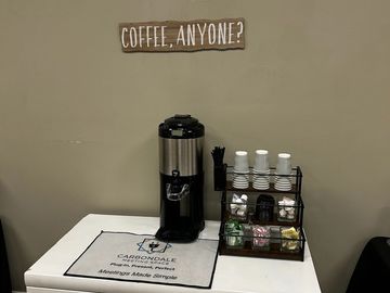 Coffee station with dispenser, cups, and condiments under a lit sign.