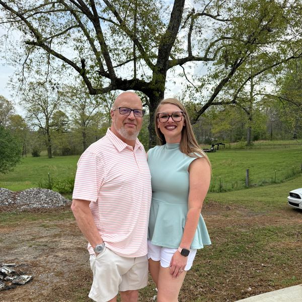 A smiling man and woman standing outdoors in front of a large tree.