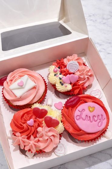 Box of four Valentine-themed cupcakes with pink and red frosting and heart decorations.