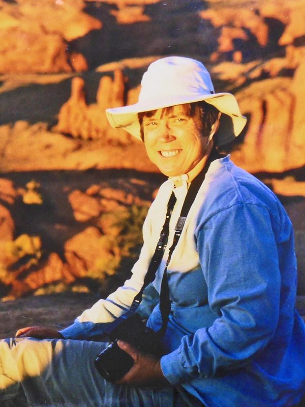 A woman wearing a white, wide-brimmed hat sits at the edge of a canyon with a camera around her neck