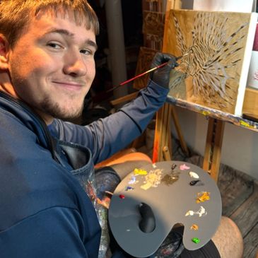 A man smiling while painting an artwork using a palette and brush.