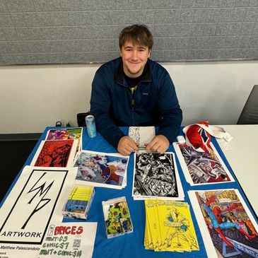 Smiling artist showcasing comic and graphic artwork at a table.