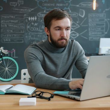 Man focused on laptop at a desk with blackboard background.