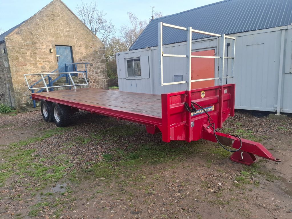 Red flatbed trailer parked outside stone and metal buildings.