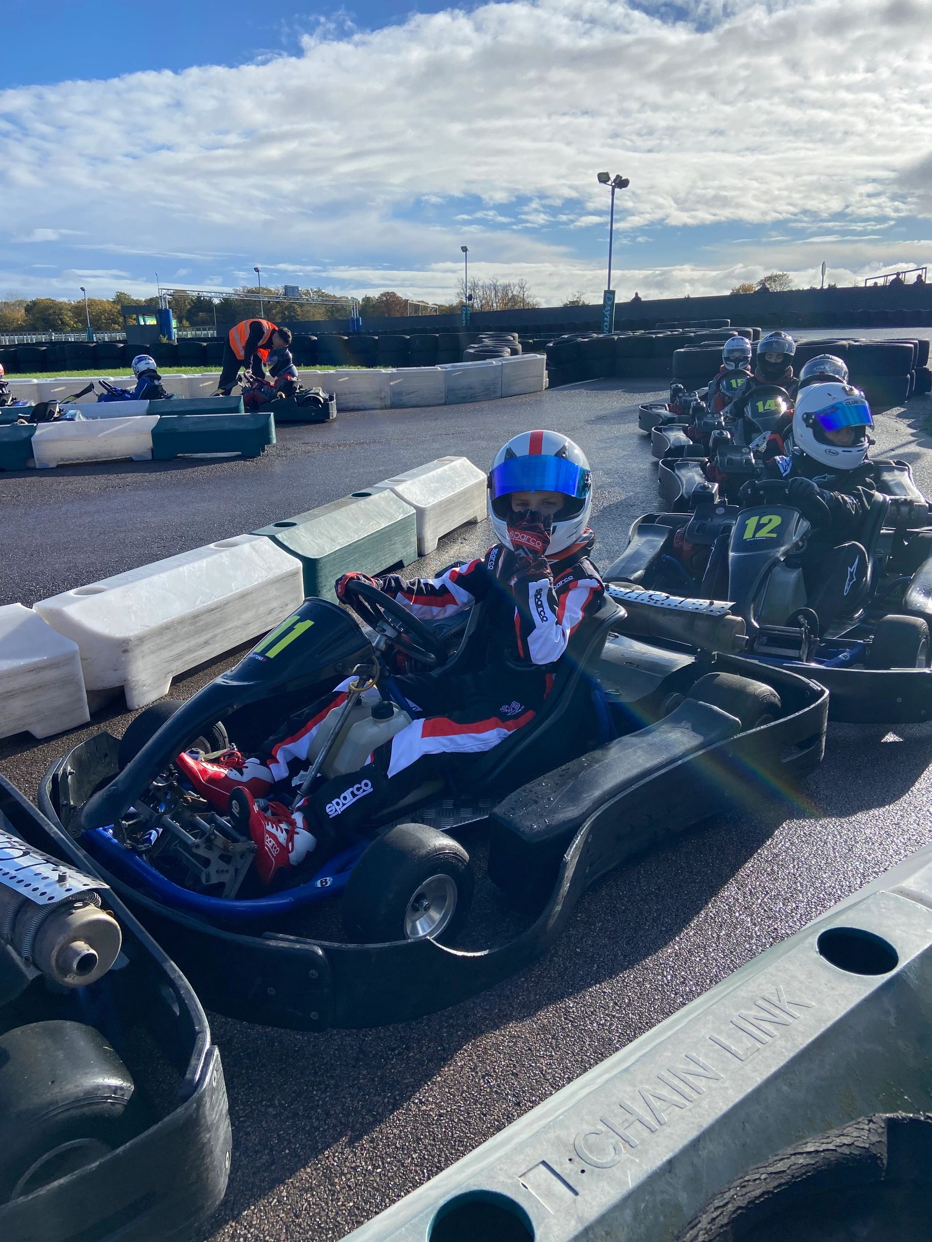 Kids in racing suits and helmets ready in go-karts on a sunny day at a track.