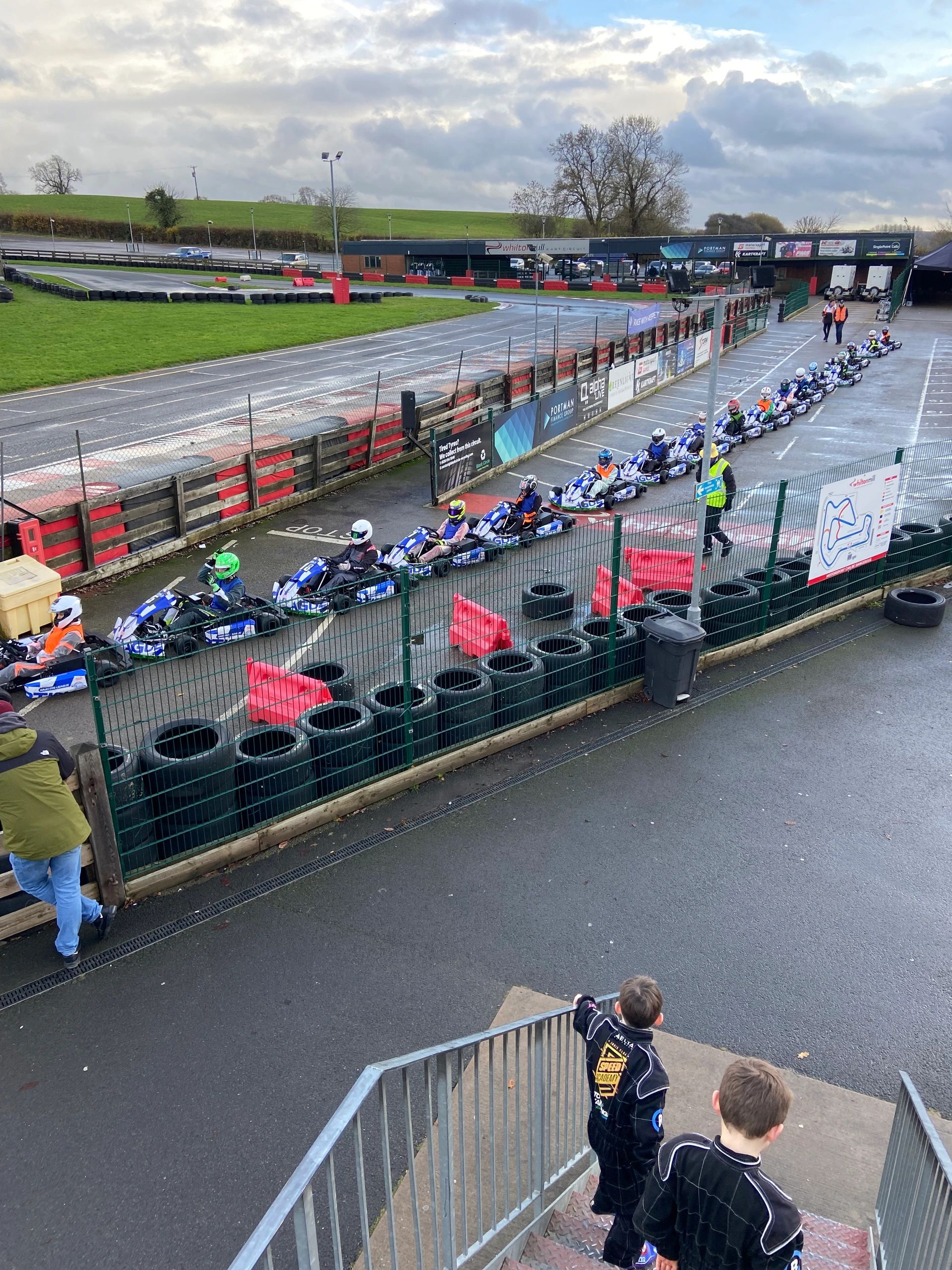 Children in racing suits watching a line of go-karts at a track.