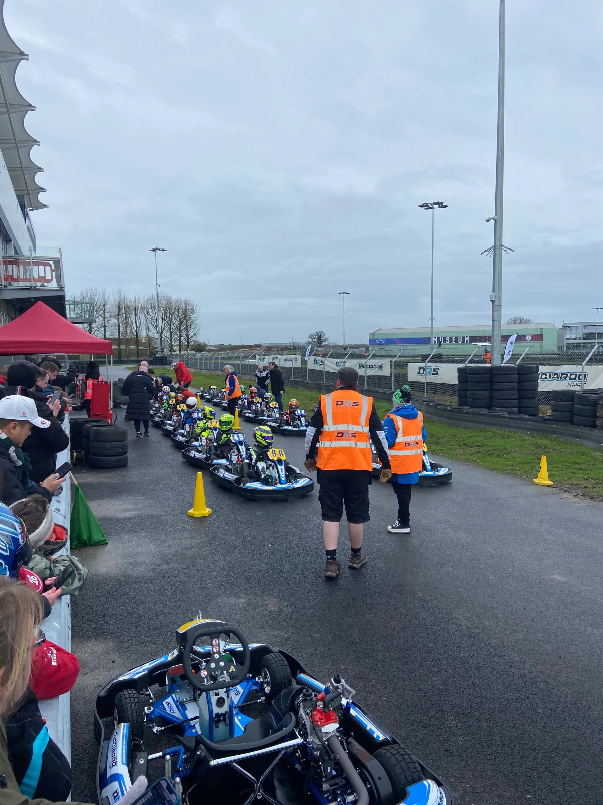 Go-kart racers lined up on a track with spectators and officials.