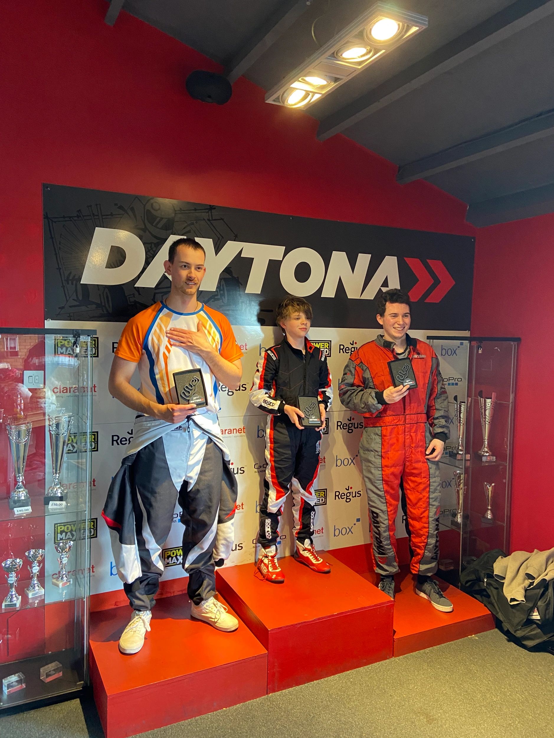 Three racers stand on a podium holding trophies at Daytona event.