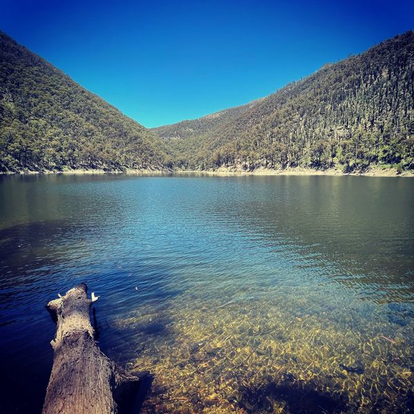 Clear lake surrounded by forested mountains under a bright blue sky.