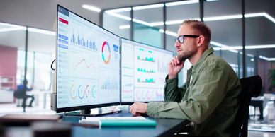 Man analyzing financial charts on dual computer monitors in a modern office.
