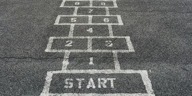 Faded hopscotch on cracked asphalt with weeds growing through.