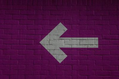 an arrow, painted in white, against a brick wall painted purple, almost magenta.  