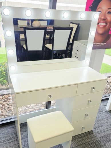 White vanity table with mirror surrounded by lights and matching stool.