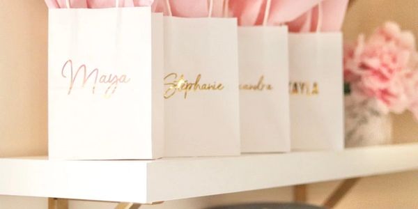 White gift bags with gold names on a white shelf, soft pink decor in background.