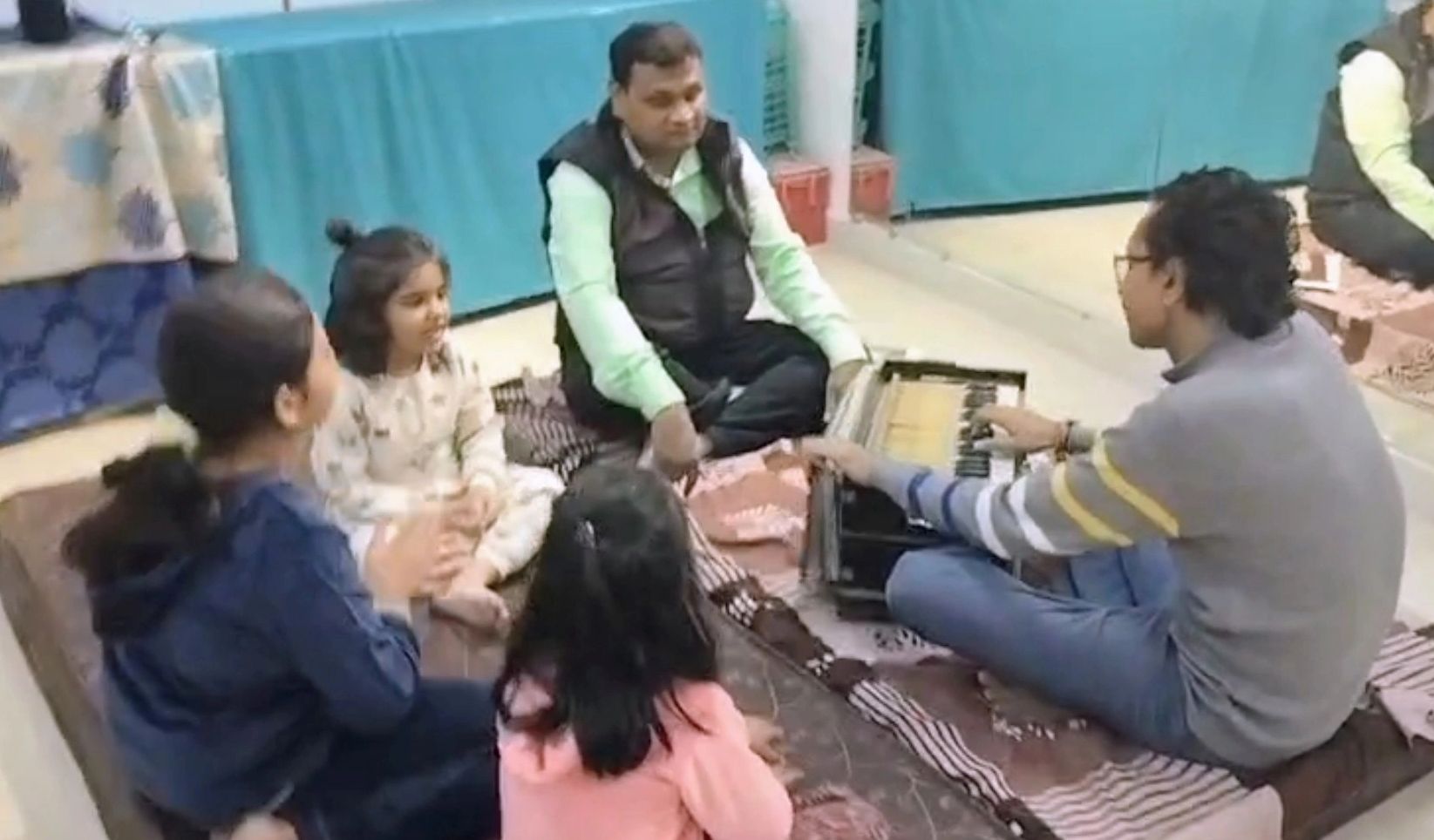 A group of people sitting on the floor, one playing a harmonium.
