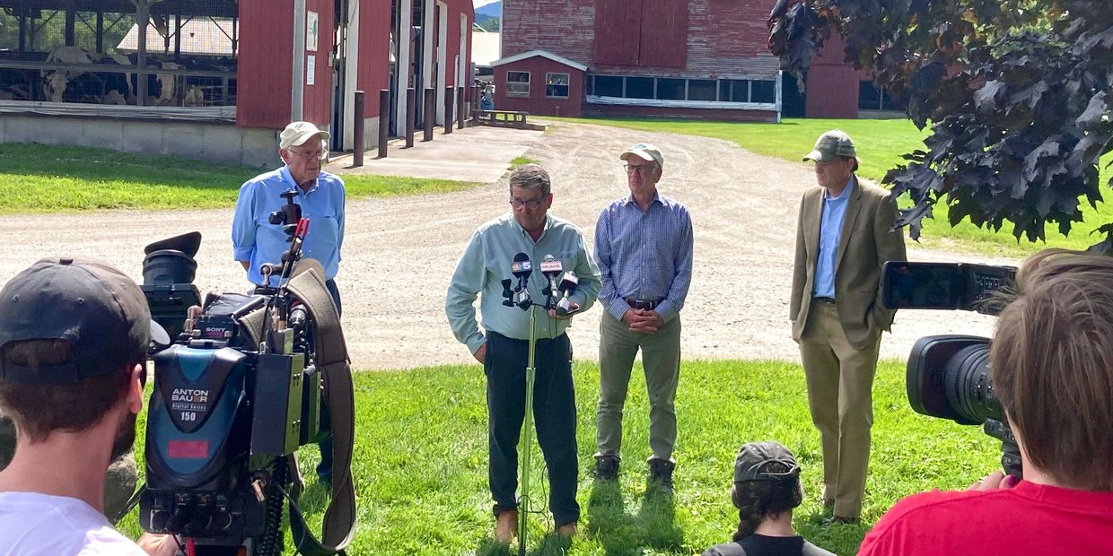 Four men speaking outdoors in front of cameras near a barn.
