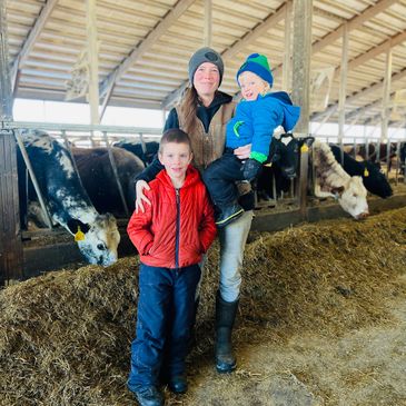 A woman and two children stand inside a barn with cows in the background.
