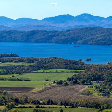 Scenic landscape with fields, forest, lake, and mountains in the background.