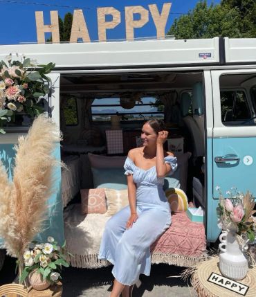Woman in light blue dress sits at van entrance with 'HAPPY' sign.