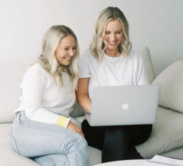 Two women smiling at a laptop on a couch.