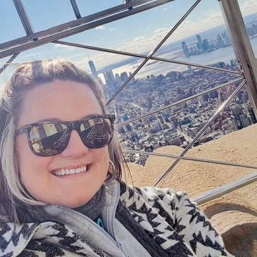 Woman smiling in sunglasses with city skyline behind her.