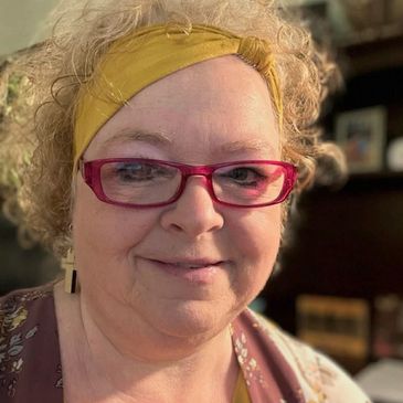A smiling woman with curly hair, red glasses, and a yellow headband.