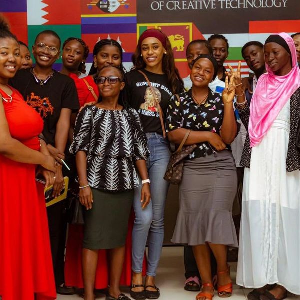A diverse group of smiling young women posing indoors with colorful flags in the background.