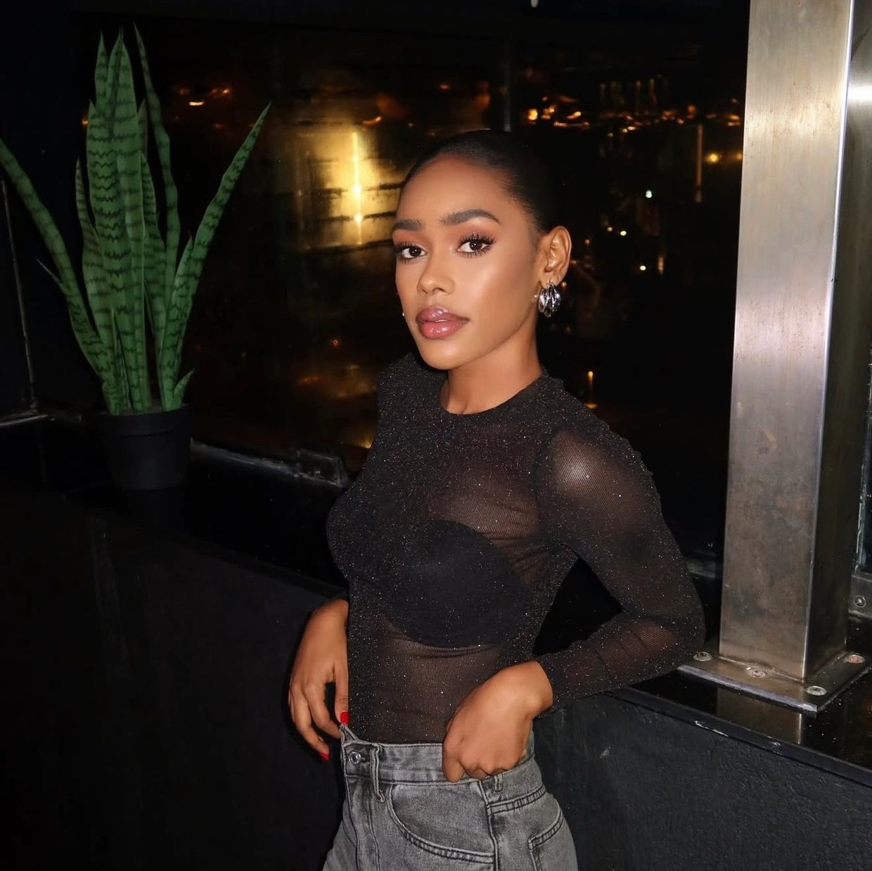 A stylish woman poses confidently in a sheer black top and high-waisted jeans at night.