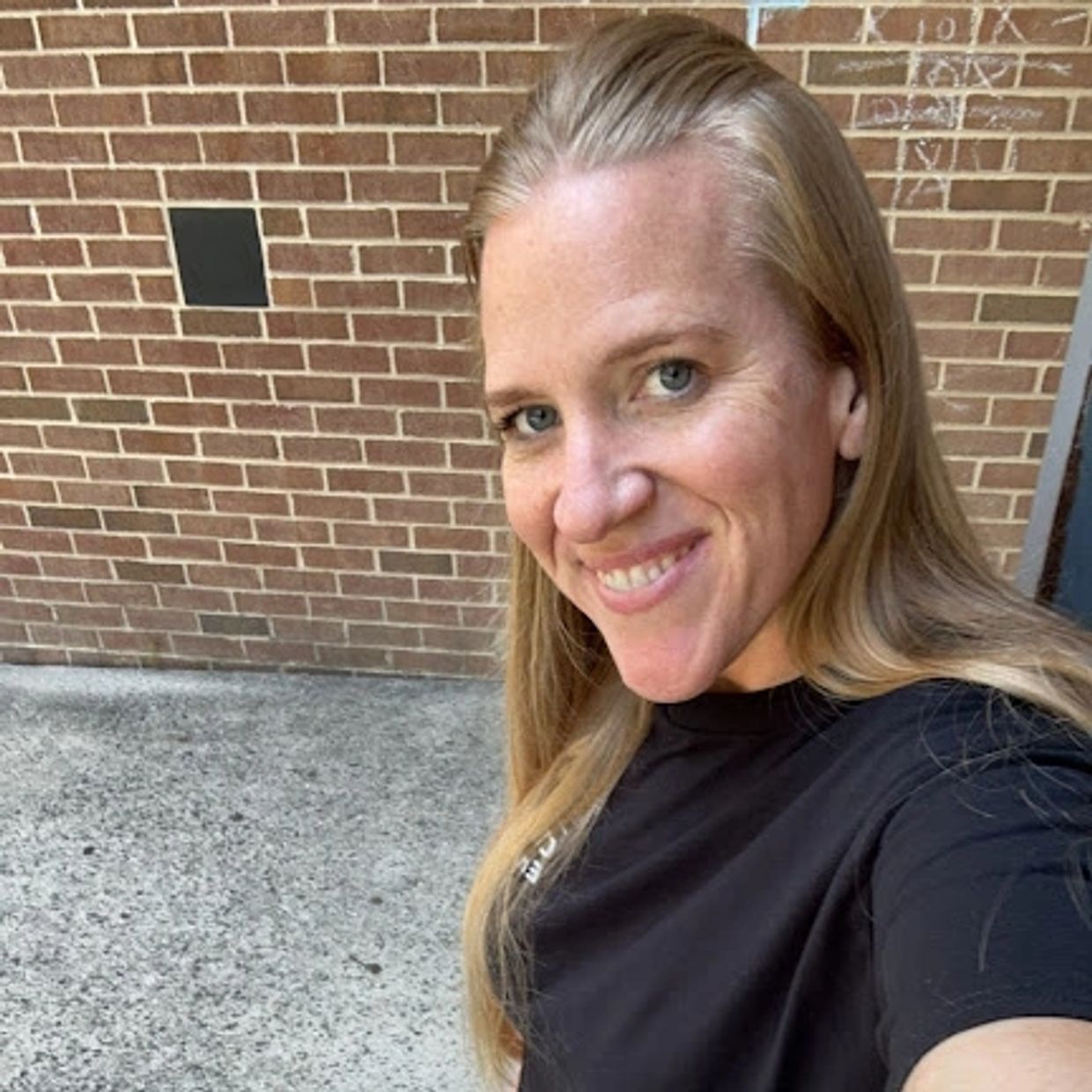 Smiling woman with long blonde hair wearing a black shirt against a brick wall.