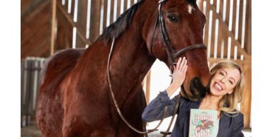 Photo of large horse and blonde woman holding Horse Girl book — Publishers Weekly Photo of the Day.