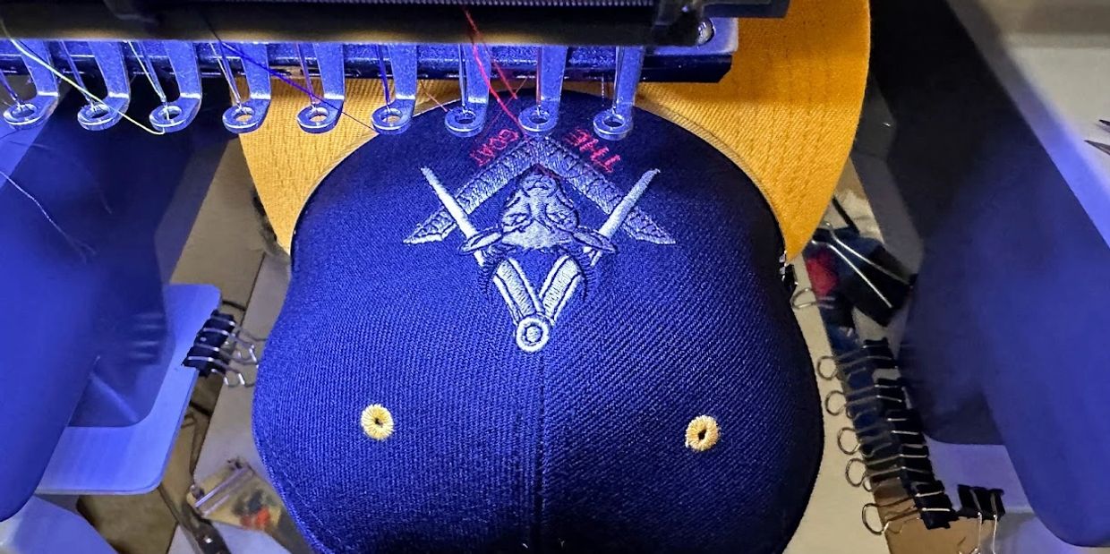 Embroidery machine stitching a detailed design on a navy and yellow cap.