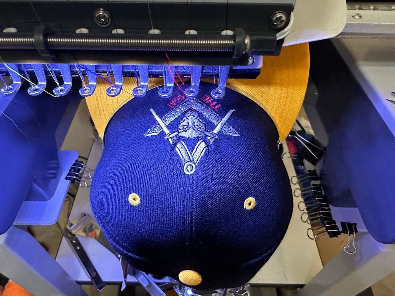 Embroidery machine stitching a detailed design on a navy and yellow cap.