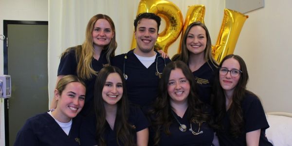 Group of seven smiling medical students in navy scrubs with stethoscopes, posing indoors.