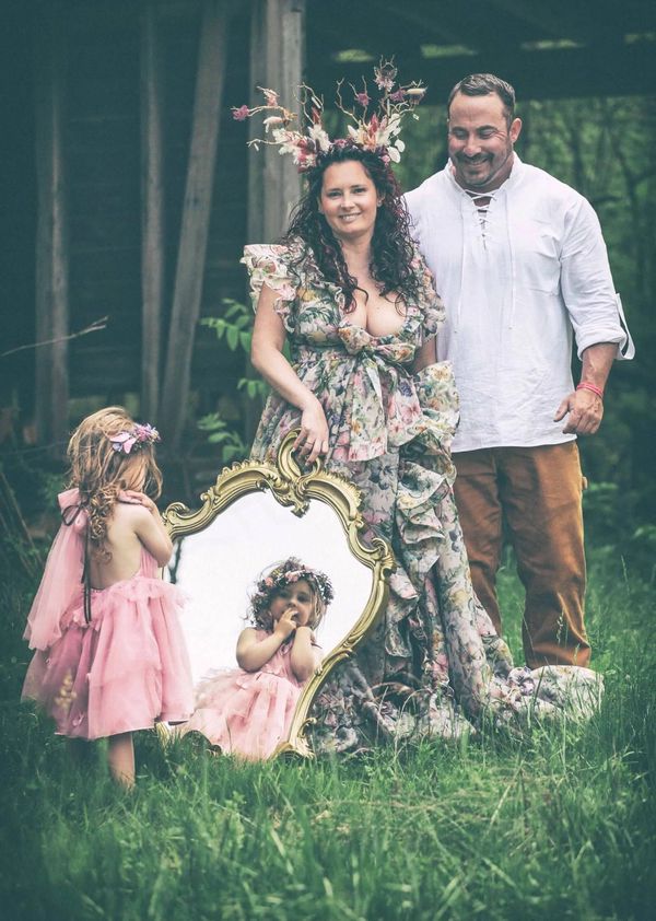 Family portrait with a child in pink dress and floral-themed adults near an ornate mirror outdoors.