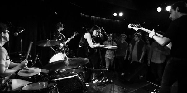 A black & white image of a band playing on stage closely surrounded by spectators