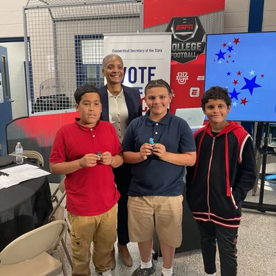 Three boys and a woman pose together in a voting-related event setting.