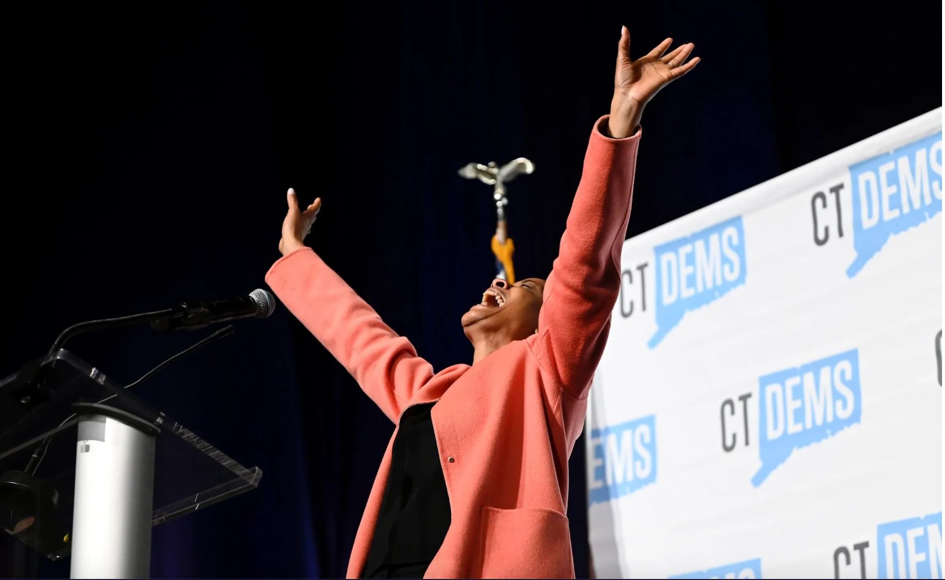 Person passionately speaking with arms raised at CT Dems event.