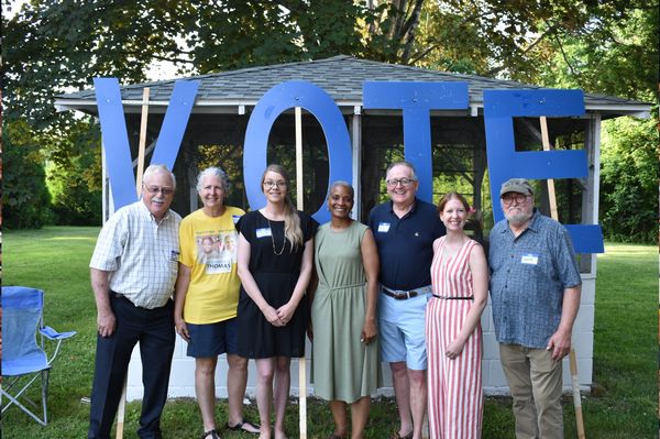 A diverse group stands together in front of large blue letters spelling VOTE outdoors.