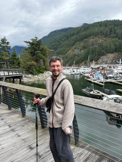 Man smiling on a wooden dock with boats and forested hills behind him.