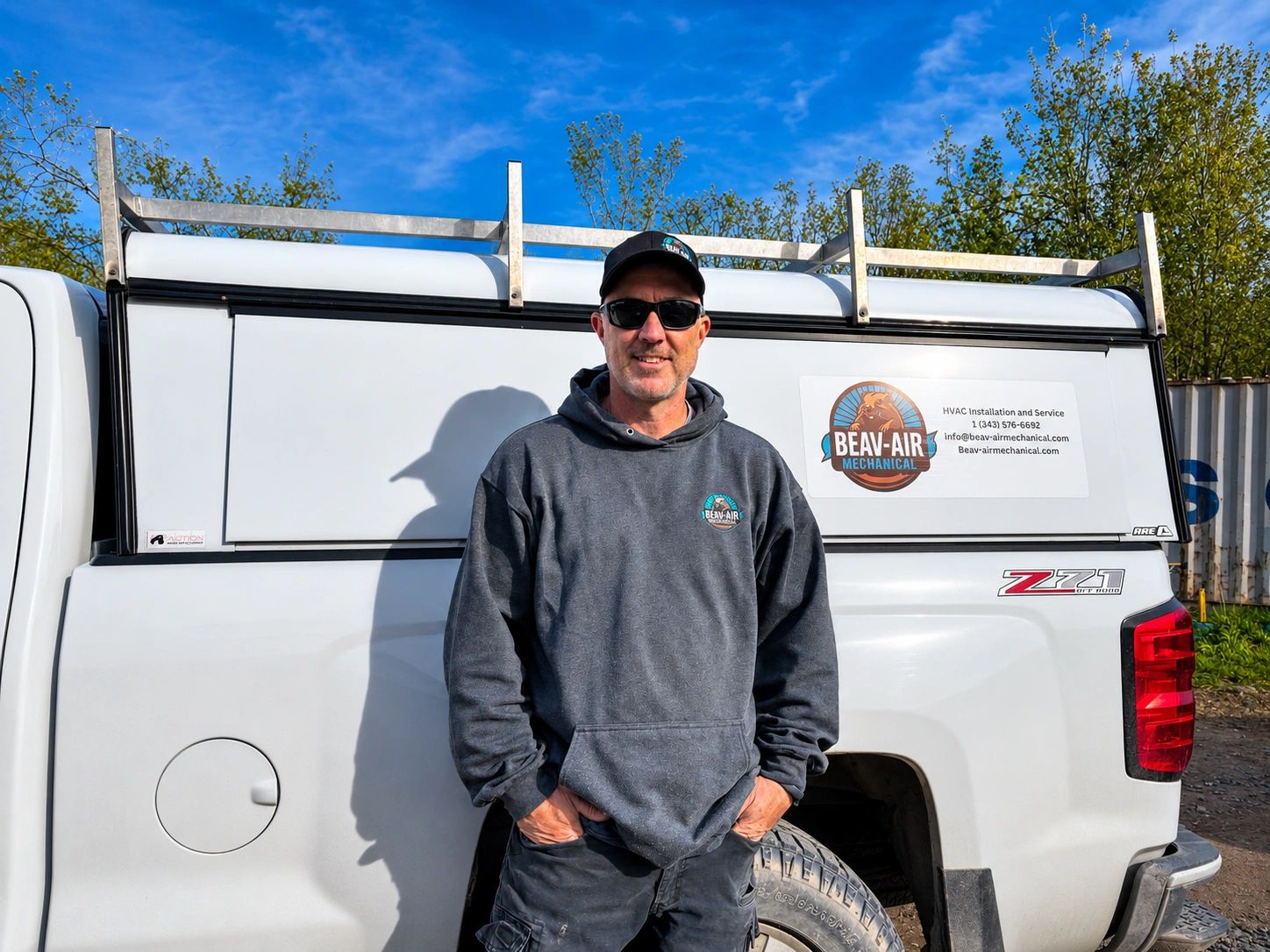 Man in sunglasses and hoodie stands by a white service truck with company logo.