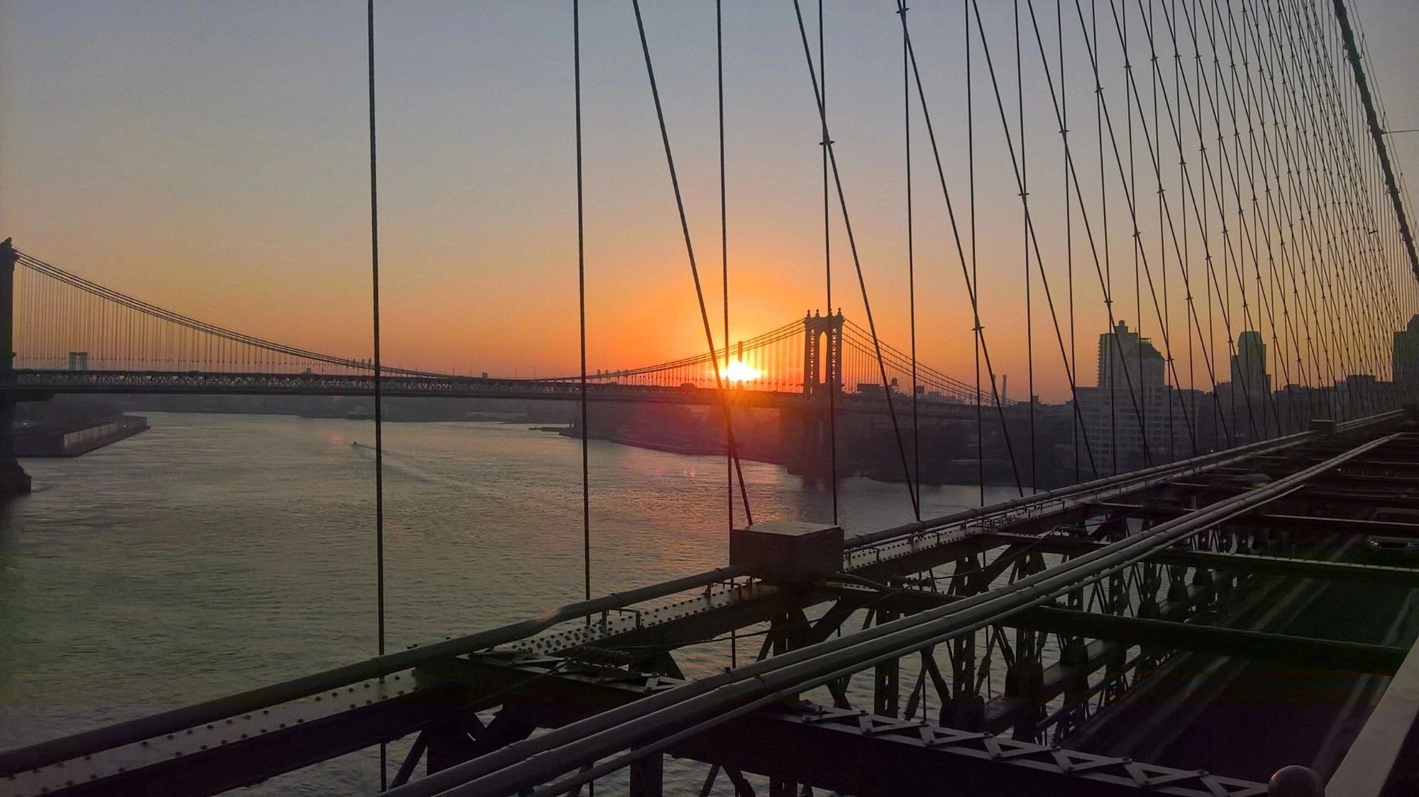 Sunset view of a suspension bridge over a river with city buildings in the background.