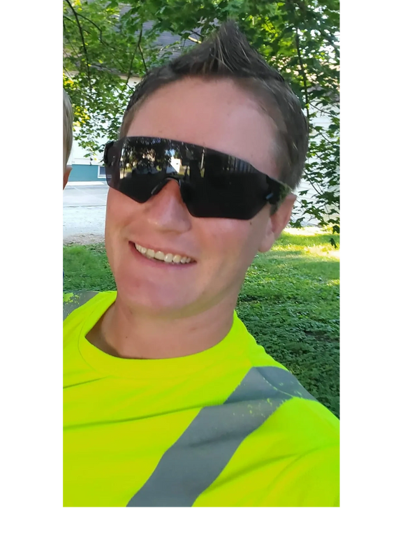 Man in neon yellow safety shirt and dark sunglasses smiling outdoors.