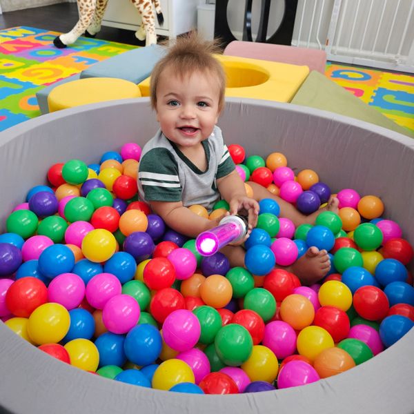 Happy toddler playing in a colorful ball pit holding a light-up toy.