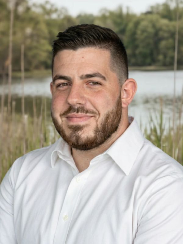 Man with beard smiling outdoors near a lake in a white shirt.