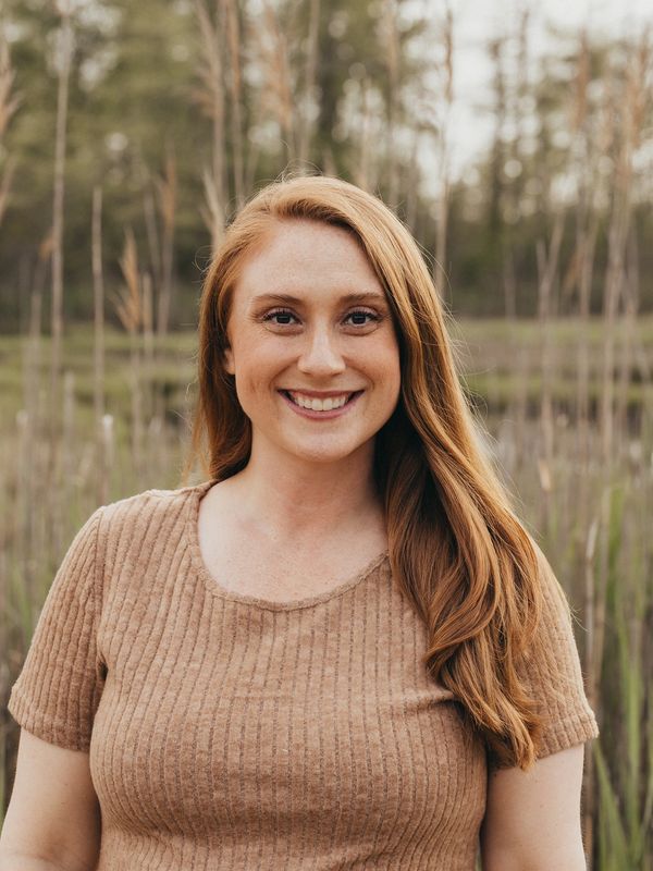 Smiling woman with long red hair in a beige top outdoors.